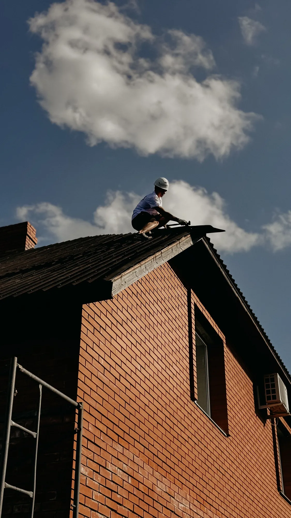Roofer in a hard hat working at the edge of a shingle roof on a brick house